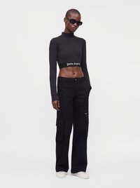 Λεπτό άτομο φοράει μαύρο crop top με μακριά μανίκια, μαύρο cargo παντελόνι, λευκά αθλητικά παπούτσια και σκούρα γυαλιά ηλίου, στέκεται μπροστά σε λευκό φόντο.