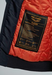 Nylon-Flugjacke mit einem orangefarbenen gesteppten Innenfutter und einem schwarzen gerippten Saum. Enthält ein Pflegeetikett mit Produktspezifikationen und Symbolen.