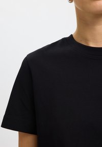 Person trägt ein schlichtes schwarzes kurzärmeliges T-Shirt mit Rundhalsausschnitt und einem kleinen Creolen-Ohrring sichtbar am linken Ohr.