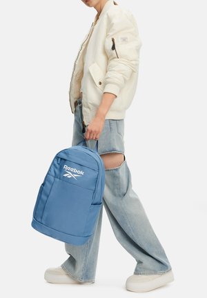 Persona con chaqueta blanca y jeans azules claros rotos sujetando una mochila azul de Reebok por la asa superior.