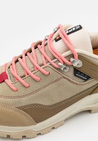 Beige wasserfester Schuh mit pinkfarbenen Schnürsenkeln, strukturiertem Obermaterial und braunen Akzenten. Verfügt über ein markantes Branding und verstärkte Ösen für Langlebigkeit.