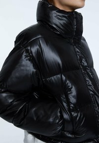 Giacca puffer nera imbottita con colletto alto, finitura lucida e sezioni a quadri orizzontali. Presenta una zip frontale e polsini elasticizzati.