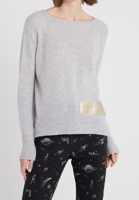 Vaaleanharmaa neule, joka on valmistettu pehmeästä kankaasta, etuosassa kultainen korostusläiskä, yhdistettynä mustiin housuihin, joissa on avaruusteema.