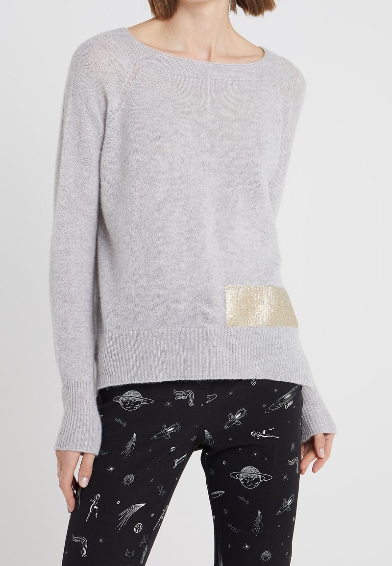 Vaaleanharmaa neule, joka on valmistettu pehmeästä kankaasta, etuosassa kultainen korostusläiskä, yhdistettynä mustiin housuihin, joissa on avaruusteema.