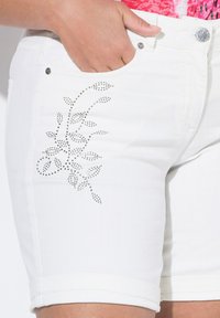 Shorts de mezclilla blanca con un ajuste estructurado, que presentan un patrón decorativo de hojas en strass plateados en el bolsillo delantero.