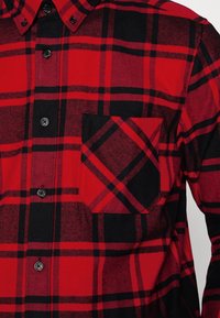 Camicia a quadri rossa e nera con tasca sul petto, dotata di bottoni neri e un motivo di tessuto testurizzato.