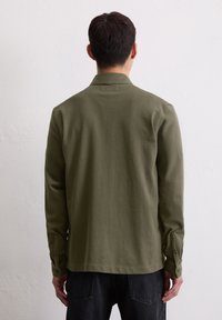 Camicia a maniche lunghe verde oliva con colletto, tessuto texturizzato e polsini con bottone, abbinata a jeans scuri.