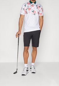 Pánská polokošile s krátkým rukávem v bílé barvě s růžovým květinovým potiskem a zeleným límečkem, kombinovaná s černými šortkami a bílými sportovními botami.