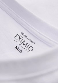 Hvid t-shirt stof med en flad, ribbet halsudskæring. Har et trykt label "EXIMIO by OMBRE," størrelse M/euro 48, og "premium kvalitet."