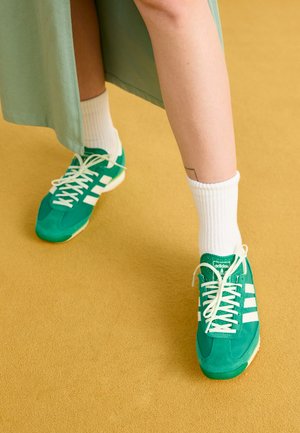 Groene sportieve schoenen met witte accenten; gemaakt van suède, ronde neus, witte veters en kenmerkende branding op de tong. Gedragen met geribbelde witte sokken.