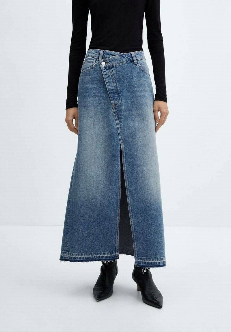 Personne portant une longue jupe en jean bleu avec une fente sur le devant, un haut noir à manches longues et des bottes noires pointues, debout devant un fond uni.