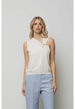 Donna con capelli biondi che indossa un top bianco asimmetrico senza maniche e pantaloni azzurri, in piedi davanti a uno sfondo grigio chiaro uniforme.