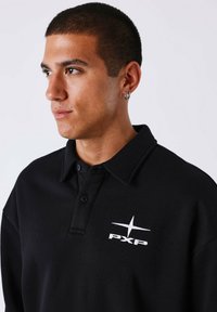 Zwarte polo met een kraag, vier knopen en een wit geborduurd logo op de voorkant. De stof lijkt glad met een zachte textuur.