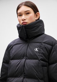 Doudoune noire rembourrée avec un col haut, présentant un design matelassé et des boutons, ornée d'un logo "CK" blanc sur la poitrine.