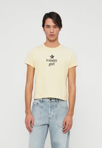Joven vistiendo una camiseta corta de color amarillo pálido con el texto "tommy girl" y un logo de estrella, combinada con unos jeans de tiro alto de color azul claro.
