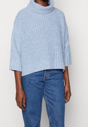 Person trägt einen hellblauen grob gestrickten Rollkragenpullover mit dreiviertel Ärmeln und mittelblauen Denim-Jeans.