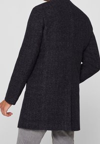 Donkergrijze wollen jas met een rechte snit, lange mouwen en een minimalistisch ontwerp. Beschikt over een achterventilatie en is mid-thigh lang.