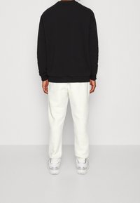 Felpa nera con collo rotondo, abbinata a pantaloni sportivi bianchi con texture e motivi intricati, e sneakers bianche con dettagli colorati.