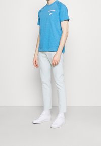 Camiseta de manga corta de color azul claro con un acabado texturizado y un pequeño logo blanco en el pecho, combinada con pantalones slim-fit de color claro y zapatillas blancas.
