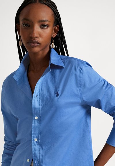 Fonott hajú nő kék gombos ingben és kagyló fülbevalóval áll egy egyszínű fehér háttér előtt.