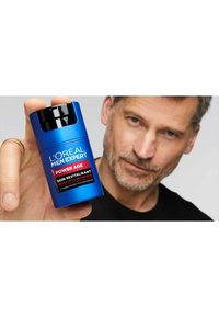 Bouteille cylindrique bleue étiquetée "L'Oréal Men Expert Power Age", avec un bouchon noir et un texte rouge. Tenue par une personne avec des cheveux courts.