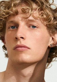 Detailný záběr osoby s krátkymi, kučeravými blond vlasmi a modrými očami, ktorá má na sebe strieborný náušník, s svetlou pleťou a viditeľnými rysmi tváre.