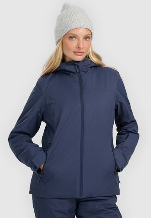 Kobieta ubrana w granatową zimową kurtkę na zamek, dopasowane spodnie oraz jasnoszary dziany beret, stoi z rękami w kieszeniach.