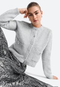 Hallide tekstuuriga hall kardigan ümarate kaelustega, ees taskud ja nööbid, koos läikiva sädelevate mustade seelikutega.