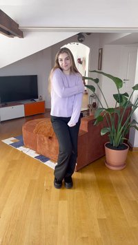 Violetinis megztinis ilgomis rankovėmis, juodos kelnės ir juodi batai. Fone matomas apvalus oranžinis sofa, žalias augalas ir medinės grindys.