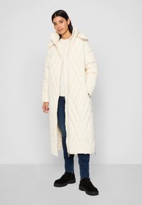 White quilted long coat with a high collar and hood, paired with a light sweater and blue jeans. Black boots complete the outfit.