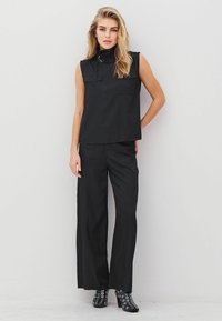 Ženska z dolgimi blond lasmi, oblečena v črno brezrokavno majico z visokim ovratnikom, široke črne hlače in črne čevlje s peto z srebrnimi okovji.