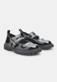 Zapatos de charol negros con punta redondeada, que cuentan con dos hebillas ajustables y una suela de goma texturizada para mayor agarre.