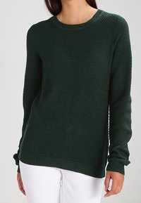 Grüner Strickpullover mit strukturierter Musterung, rundem Ausschnitt sowie gerippten Bündchen und Saum, kombiniert mit weißen Hosen.