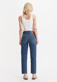 Pantalones vaqueros de mezclilla azul de tiro alto con pierna recta, bolsillos traseros visibles y una etiqueta de cuero en la cintura. Llevados con una camiseta sin mangas blanca.