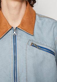 Ljusblå jacka med en beige läderkrage, har en framzip och en ficka på bröstet med blixtlås, med blå accenter och kontrasterande sömmar.