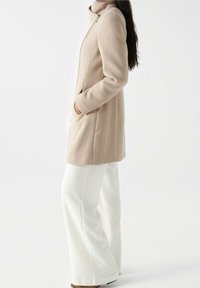 Beige villakangas takki, jossa on korkea kaulus ja sivutaskut, päällä valkoiset leveälahkeiset housut. Silkkinen pinta ja minimalistinen muotoilu.