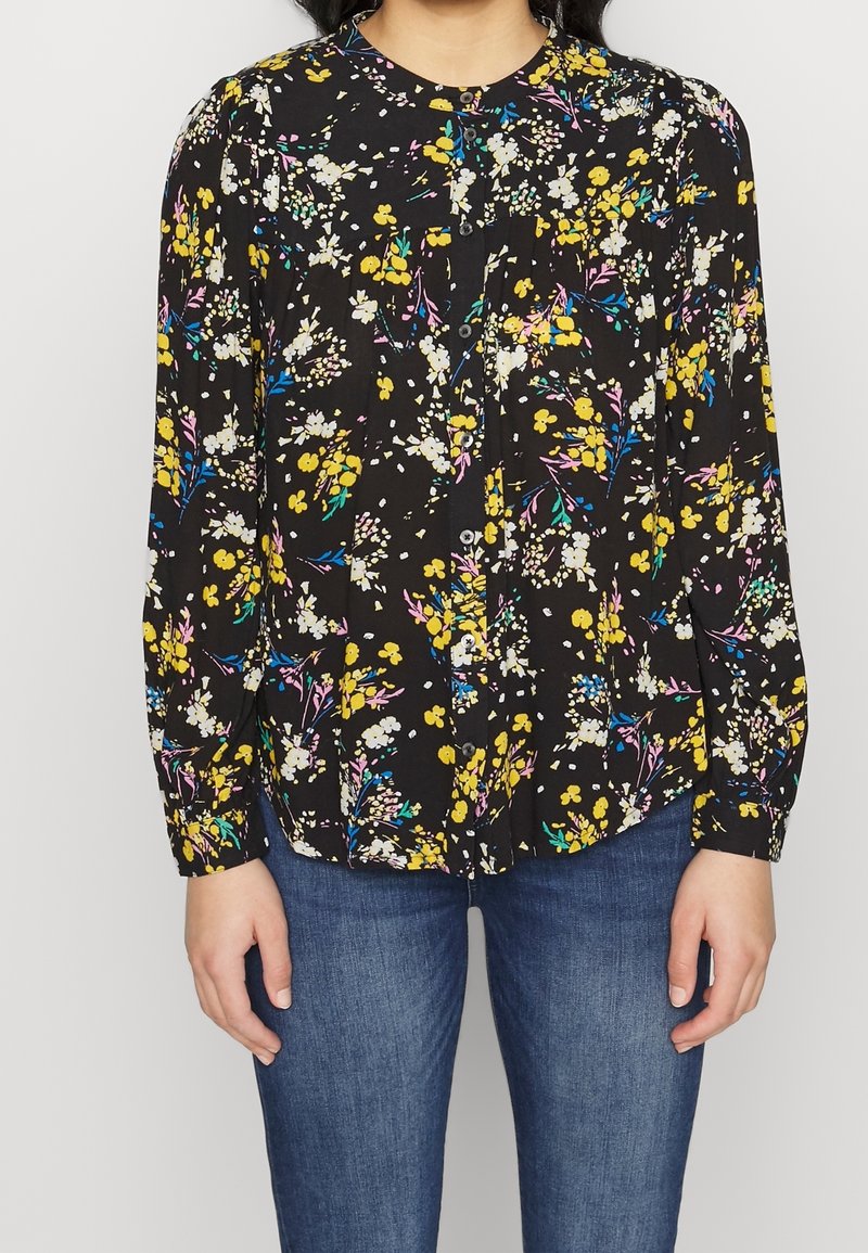 Femme portant un chemisier noir à manches longues boutonné avec un motif floral multicolore et un jean bleu, debout devant un fond uni.