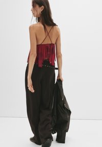 Una donna in pantaloni neri larghi e un top rosso con spalline, che tiene una giacca di pelle nera, in piedi contro uno sfondo bianco semplice.