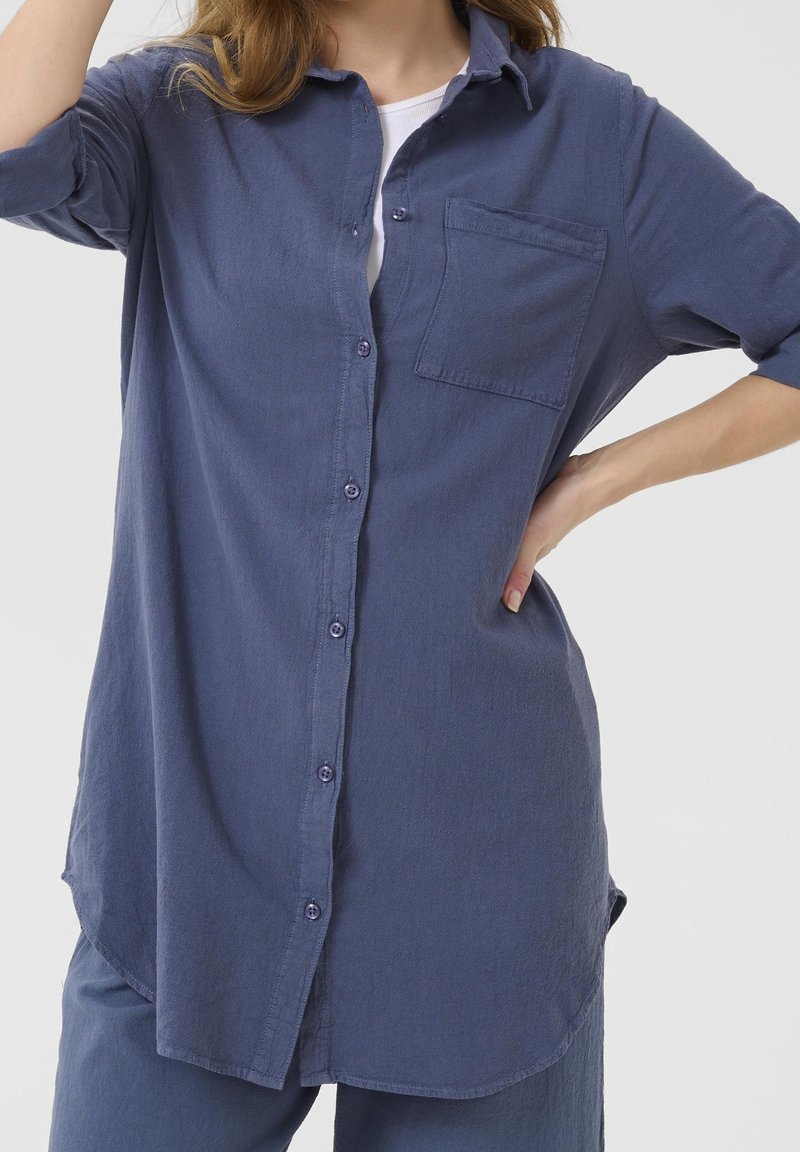 Mujer vestida con una camisa azul de manga larga y botones, con un bolsillo en el pecho, sobre una camiseta blanca. Tiene una mano en la cadera y la imagen está recortada a la altura del cuello y las rodillas.