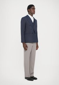 Tengerészeti dupla mellű zakó fekete gombokkal, szürke szabott nadrággal és fekete cipővel. A zakó alatt fehér ing látható.