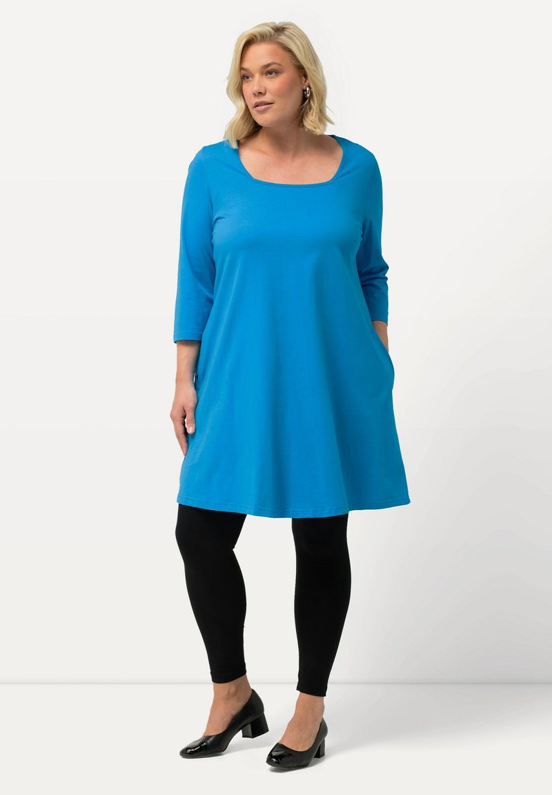 Blaues A-Linien-Kleid mit quadratischem Ausschnitt und Dreiviertelärmeln, kombiniert mit schwarzen Leggings und schwarzen High Heels. Verfügt über Seitentaschen.