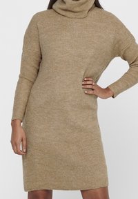 Abito a collo alto lavorato a maglia in beige, caratterizzato da una vestibilità comoda, maniche lunghe e texture a coste. L'orlo è leggermente sopra il ginocchio.