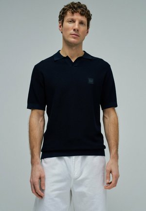 Hombre con cabello rizado que lleva una camisa polo negra de manga corta y pantalones blancos sobre un fondo gris claro.