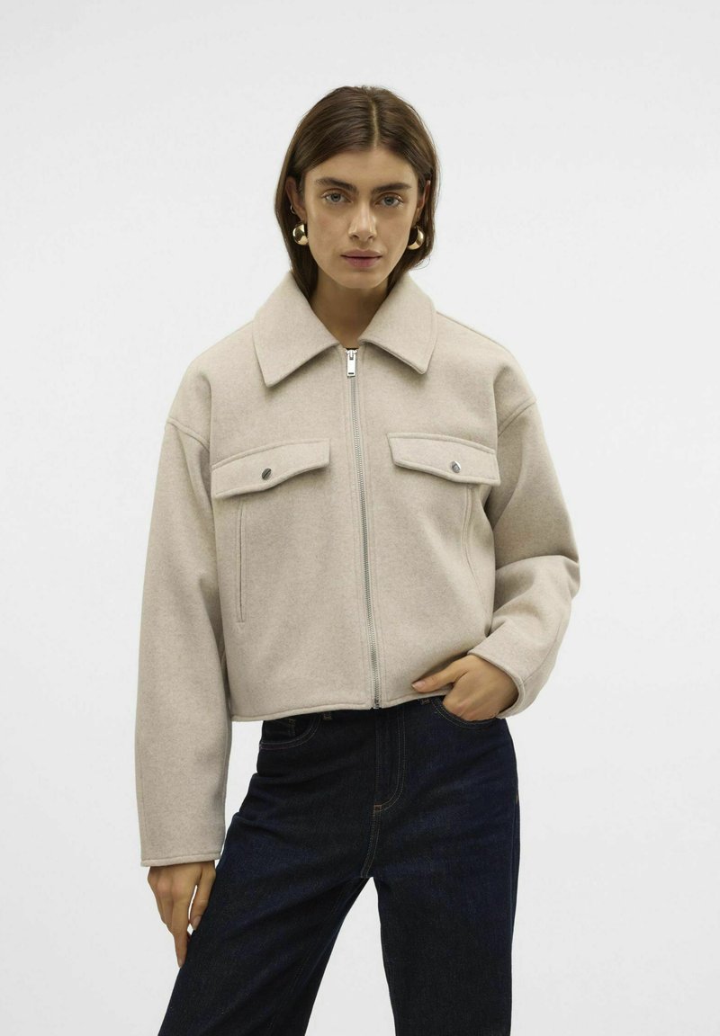 Beige zkrácená bunda vyrobená z měkké tkaniny, která má špičatý límec, zapínání na zip a dva přední kapsy se zacvakávacími knoflíky.