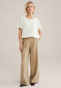 Blouse crème légère, plissée, à manches courtes, associée à un pantalon large à taille haute en beige texturé et des chaussures foncées.