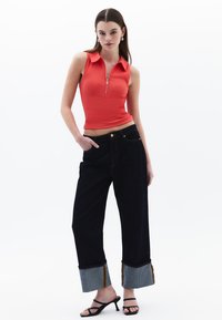 Rode mouwloze top met een kraag en ritssluiting, gecombineerd met donkerblauwe brede jeans met contrasterende zoom en gele stiksels.