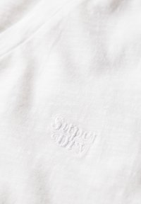 Bílá bavlněná tkanina s jemnou texturou, na které je vyšitý logo "Superdry" ve shodné bílé nitě. Jednoduchý, minimalisticý design.