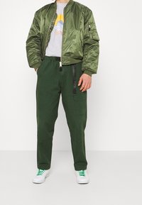Uomo che indossa una giacca bomber verde oliva, una t-shirt grigia con grafica, pantaloni cargo verde scuro e sneakers bianche con lacci verdi, in piedi contro uno sfondo bianco.