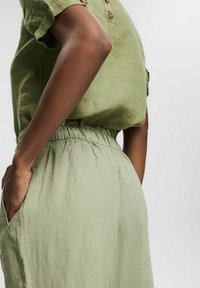 Persona que lleva pantalones de lino verde claro con cintura elástica y una camiseta de manga corta a juego, con la mano en el bolsillo, frente a un fondo liso.