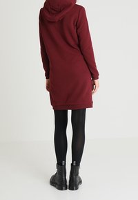Vestido sudadera burdeos, de longitud a la rodilla, con capucha y dobladillo acanalado. Combinado con mallas negras y botines. Tejido suave y texturizado.
